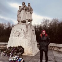 Osoba stojí pri pamätníku na ceste, oblečená v čiernej bunde s červenými vlasmi, v pozadí je obloha a stromy. Pamätník na ceste s osobou v čiernej bunda a červenými vlasmi.