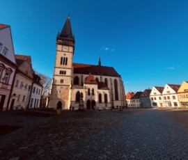 Historický kostol s vysokou vežou na námestí, obklopený farebnými domami, za jasného dňa.