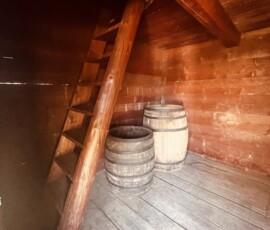 Hradná podzemná miestnosť s sudmi a schodiskom, drevené steny a schodisko, historická atmosfér.
