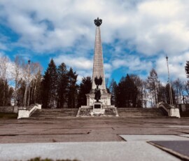 Pamätník na Mierovom námestí v Šuhajovciach s modrou oblohou a stromami v pozadí.