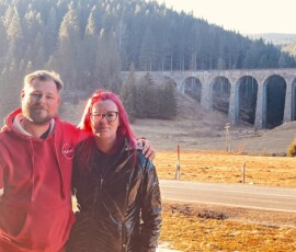 Fotografia dvojice na výlete v prírode s historickým viaduktom v pozadí, zachytávajúca ich zážitky počas cesty. Šuhajovci na cestách: dvojica na výlete pri moste v prírode, s krásnym výhľadom na lesy a historický.