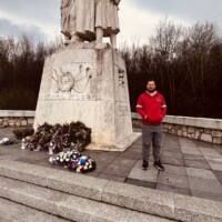 Muž stojí pri pamätníku Šuhajovci, obklopený kvetmi, na ceste v obci Šuhajovci, Slovensko. Pamätník Šuhajovci s mužom v červenom tričku v popredí.