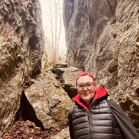 Žena s červenými vlasmi a čiernou bundou stojí medzi skalami v prírodnom prostredí počas výletu v Šuhajovcoch. Žena v prírode medzi skalami počas výletu v Šuhajovcoch.
